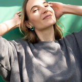 A woman with shoulder-length hair wearing a loose gray top stands in sunlight against a green background, smiling slightly with her hands raised near her head, showcasing Minimalist Circle Stud Earrings that are hypoallergenic.