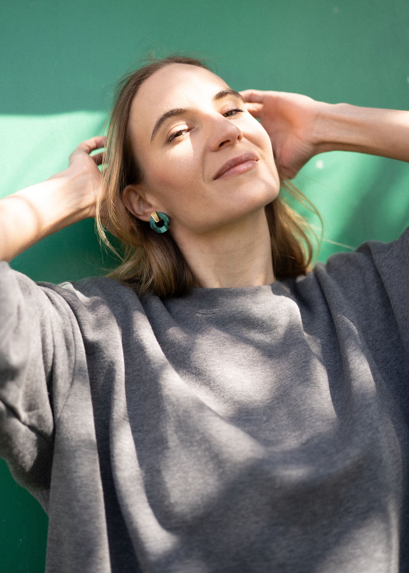 A woman with shoulder-length hair wearing a loose gray top stands in sunlight against a green background, smiling slightly with her hands raised near her head, showcasing Minimalist Circle Stud Earrings that are hypoallergenic.