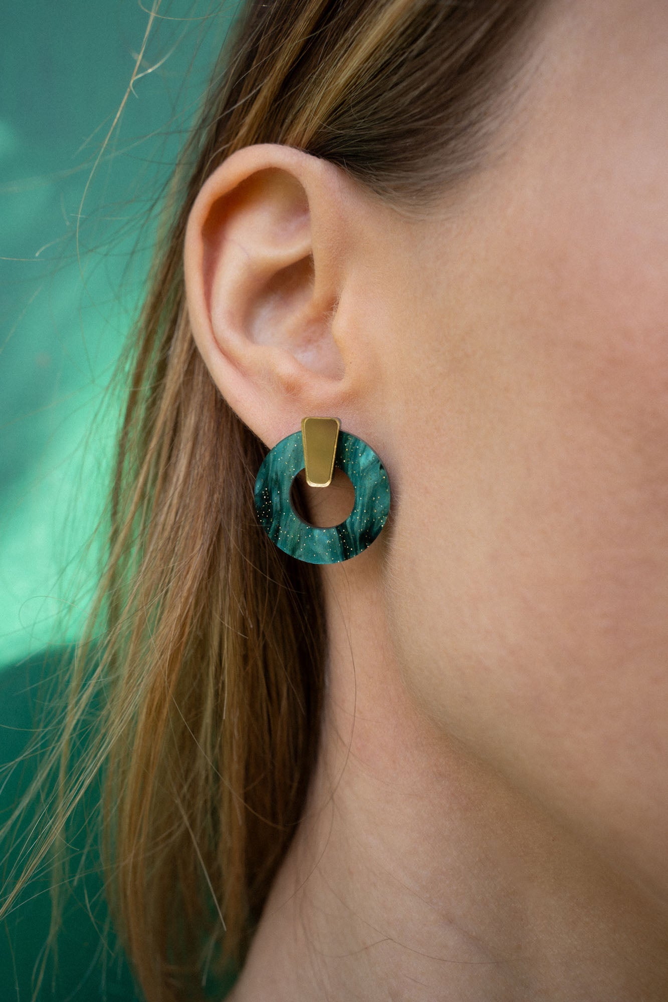 Close-up of a womans ear wearing a hypoallergenic, dark green marbled Minimalist Circle Stud Earring with a gold rectangular accent; her light brown hair partially frames her ear.