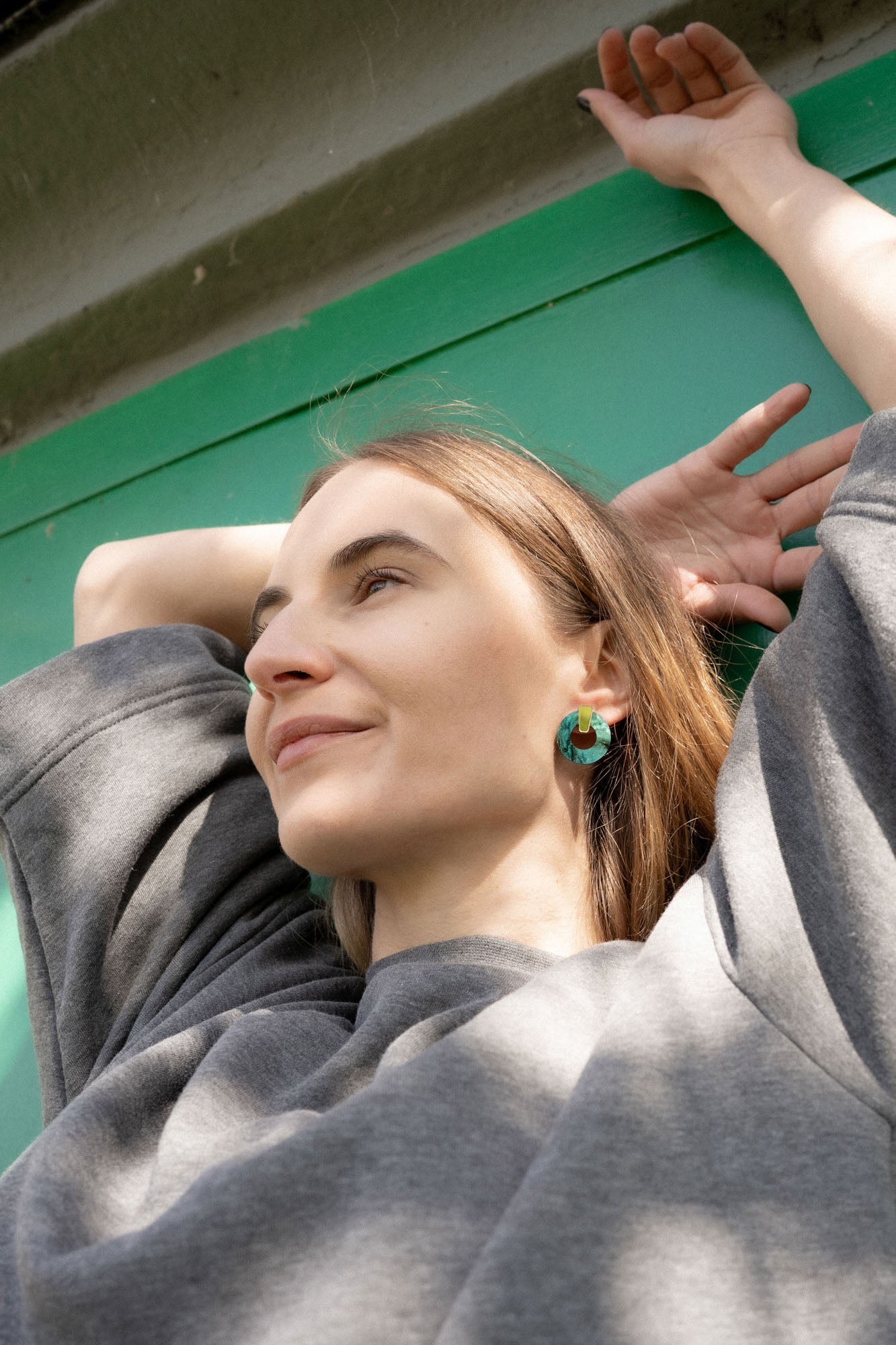 A woman in a grey sweatshirt leans back with her arms raised behind her head, smiling slightly. She stands before a green wall, sunlight on her face, and wears hypoallergenic Minimalist Circle Stud Earrings in green acrylic glass.