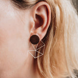 Close-up of a person with short blonde hair wearing a geometric earring featuring a dark circular stud and a wireframe cube, bringing minimalist elegance to contemporary looks.