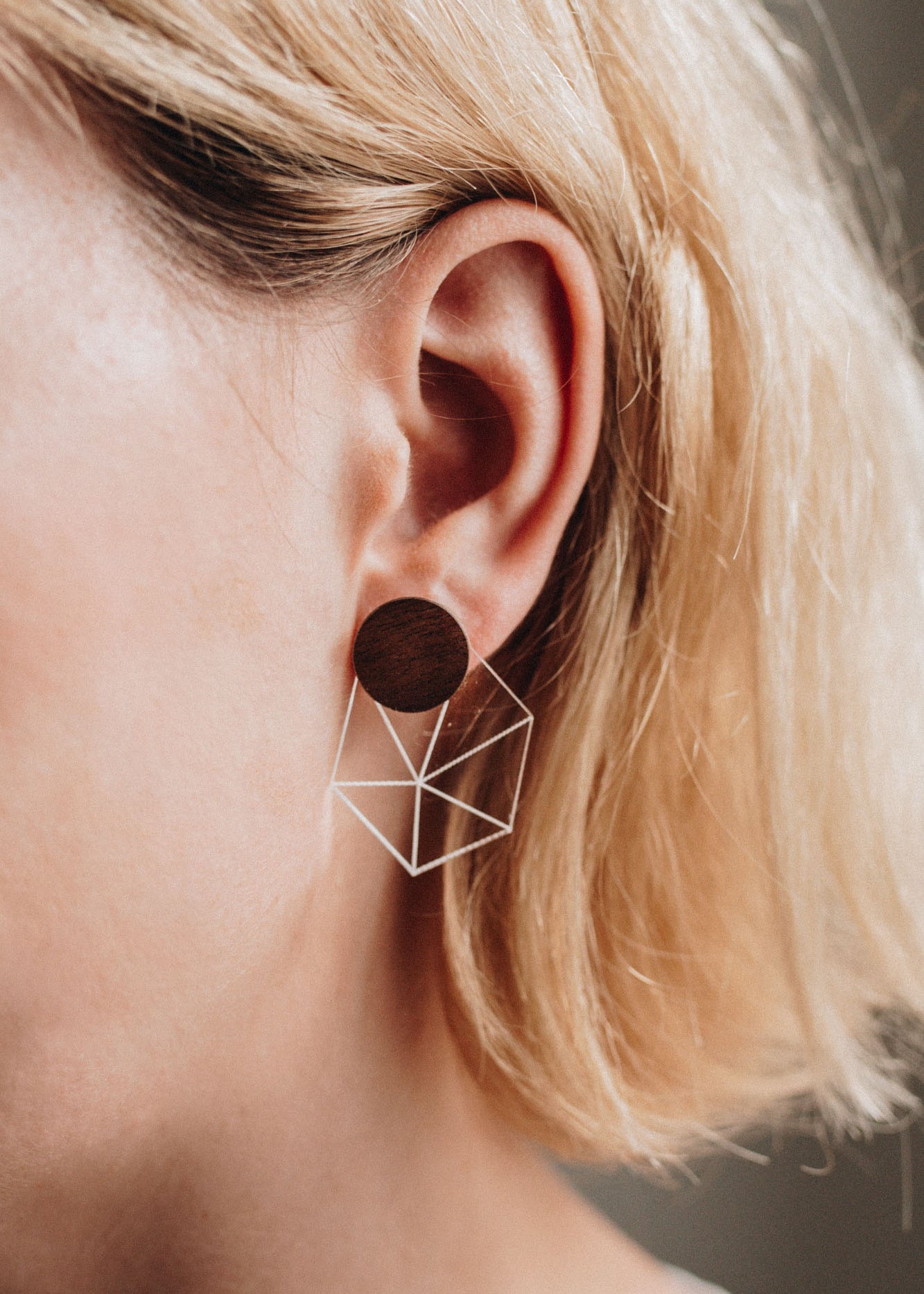 Close-up of a person with short blonde hair wearing a geometric earring featuring a dark circular stud and a wireframe cube, bringing minimalist elegance to contemporary looks.