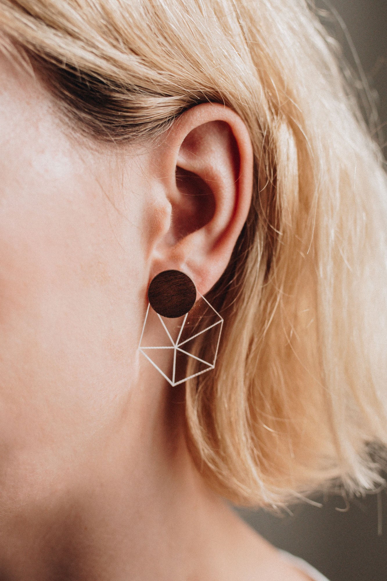 Close-up of a person with short blonde hair wearing a geometric earring featuring a dark circular stud and a wireframe cube, bringing minimalist elegance to contemporary looks.