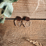 A pair of geometric earrings with wooden circular tops and silver wire shapes below, displayed on a rustic wooden surface with green leaves and dried wheat in the corners, exuding minimalist elegance.