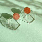 A pair of modern earrings featuring circular wooden tops attached to transparent geometric cube bases, displayed on a mint green background with soft shadows, embodying minimalist elegance.