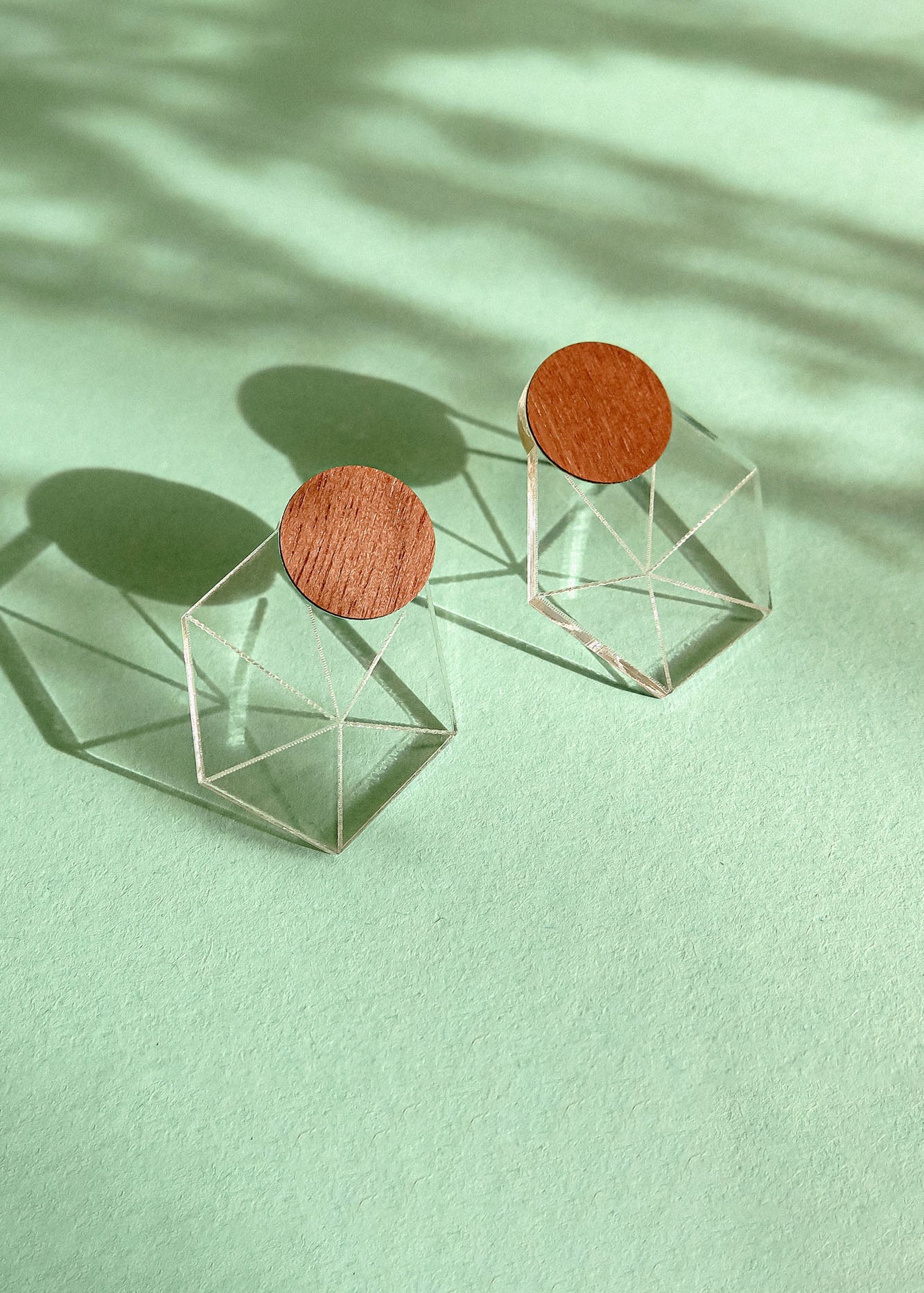 A pair of modern earrings featuring circular wooden tops attached to transparent geometric cube bases, displayed on a mint green background with soft shadows, embodying minimalist elegance.