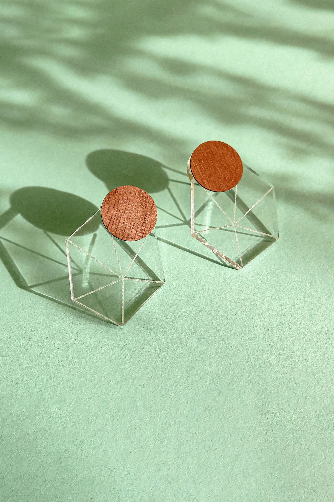 A pair of modern earrings featuring circular wooden tops attached to transparent geometric cube bases, displayed on a mint green background with soft shadows, embodying minimalist elegance.