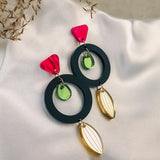 A pair of bold, modern dangle earrings with pink triangular studs, large black hoops, small green circular accents, and gold leaf-shaped dangles crafted from recycled acrylic, displayed on white fabric with decorative dried grass nearby.