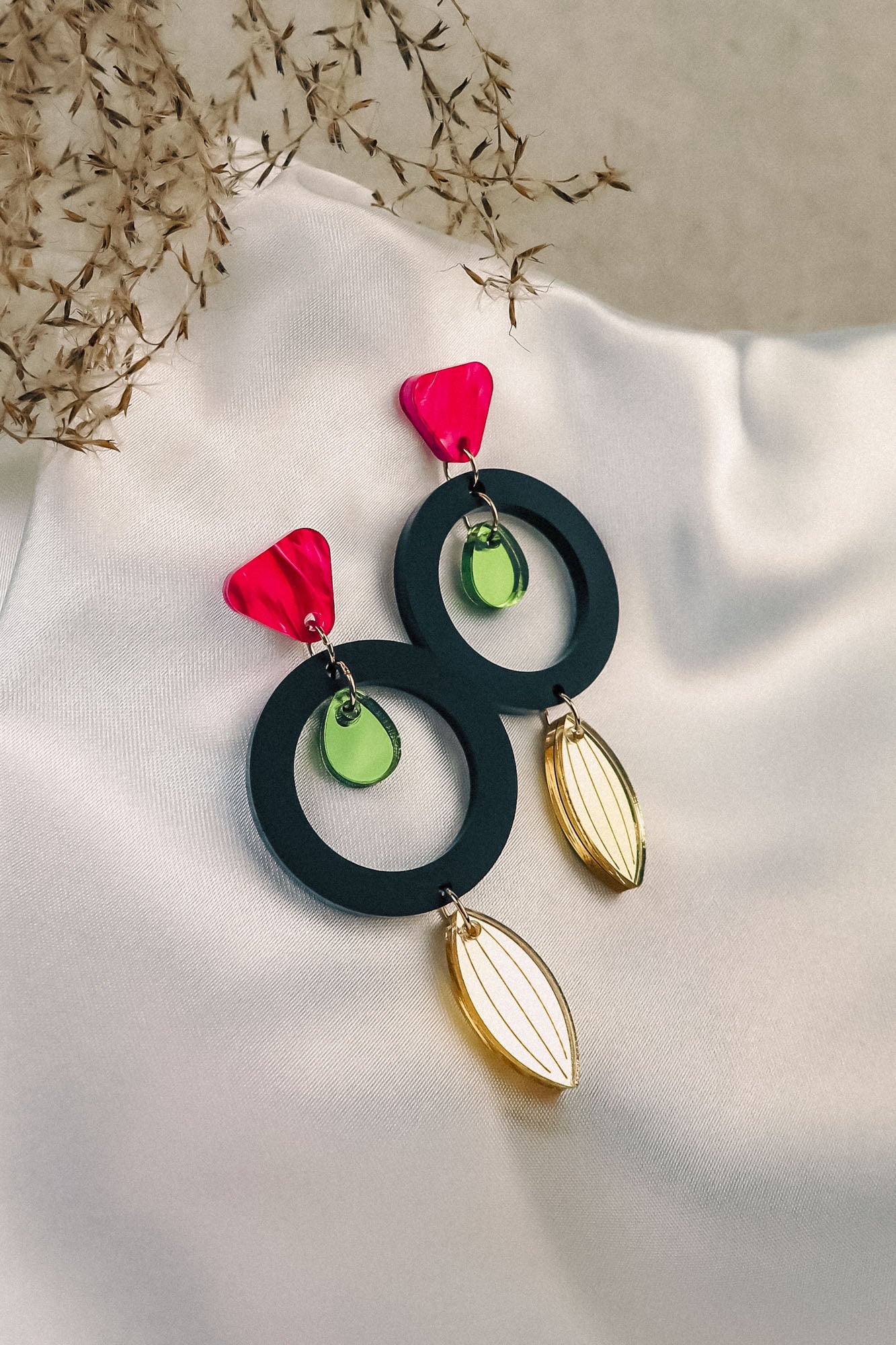 A pair of bold, modern dangle earrings with pink triangular studs, large black hoops, small green circular accents, and gold leaf-shaped dangles crafted from recycled acrylic, displayed on white fabric with decorative dried grass nearby.