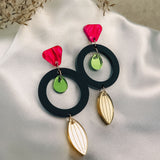 A pair of modern dangle earrings with pink geometric tops, black circles, small green discs in the center, and gold leaf-shaped pendants crafted from recycled acrylic, displayed on white satin fabric with dried foliage in the background.