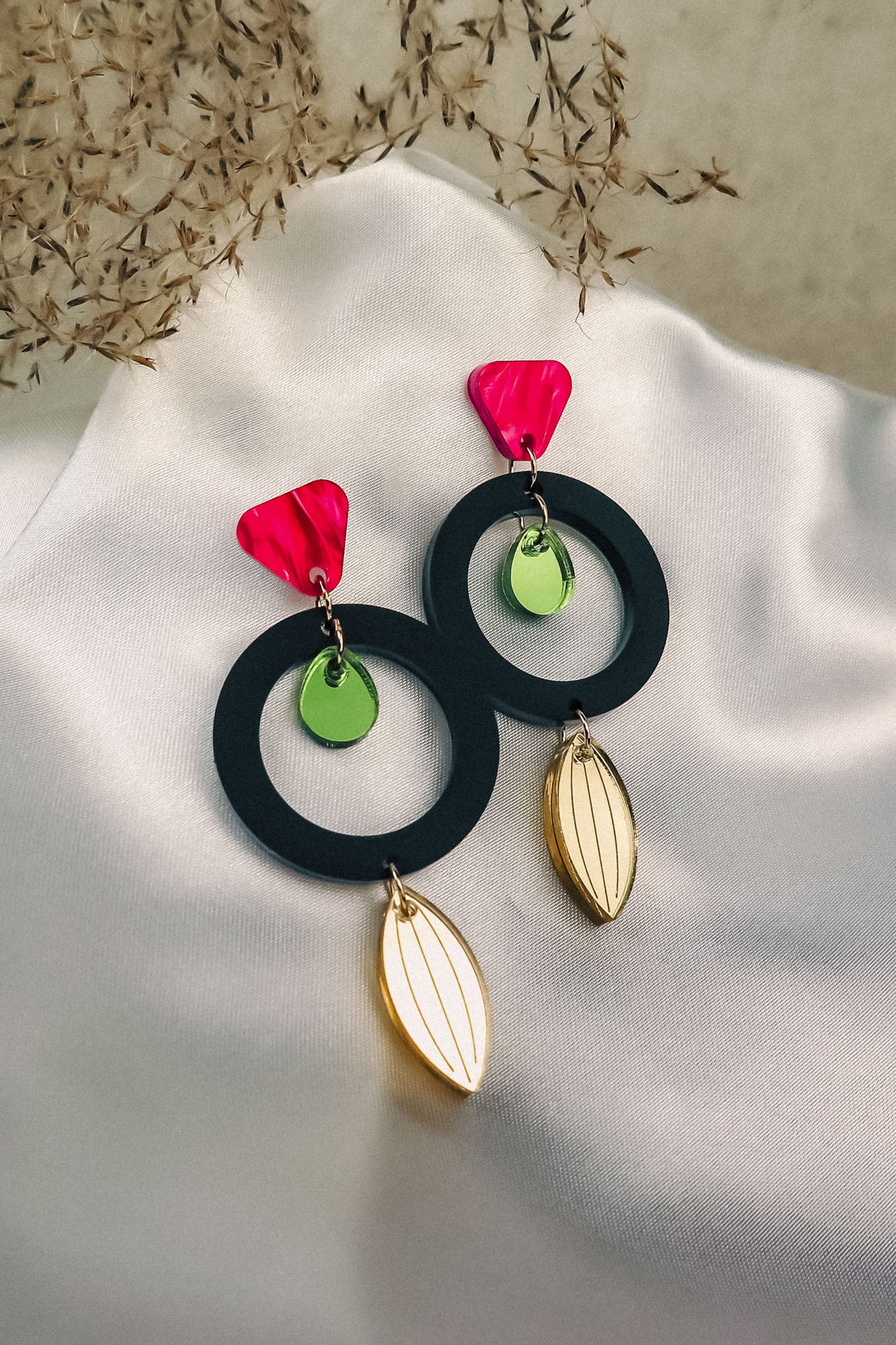 A pair of modern dangle earrings with pink geometric tops, black circles, small green discs in the center, and gold leaf-shaped pendants crafted from recycled acrylic, displayed on white satin fabric with dried foliage in the background.