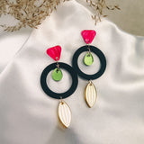 A pair of modern, colorful dangle earrings crafted from recycled acrylic, featuring pink triangle studs, green circular accents, black hoops, and gold leaf-shaped dangles displayed on white satin with dried foliage in the background.