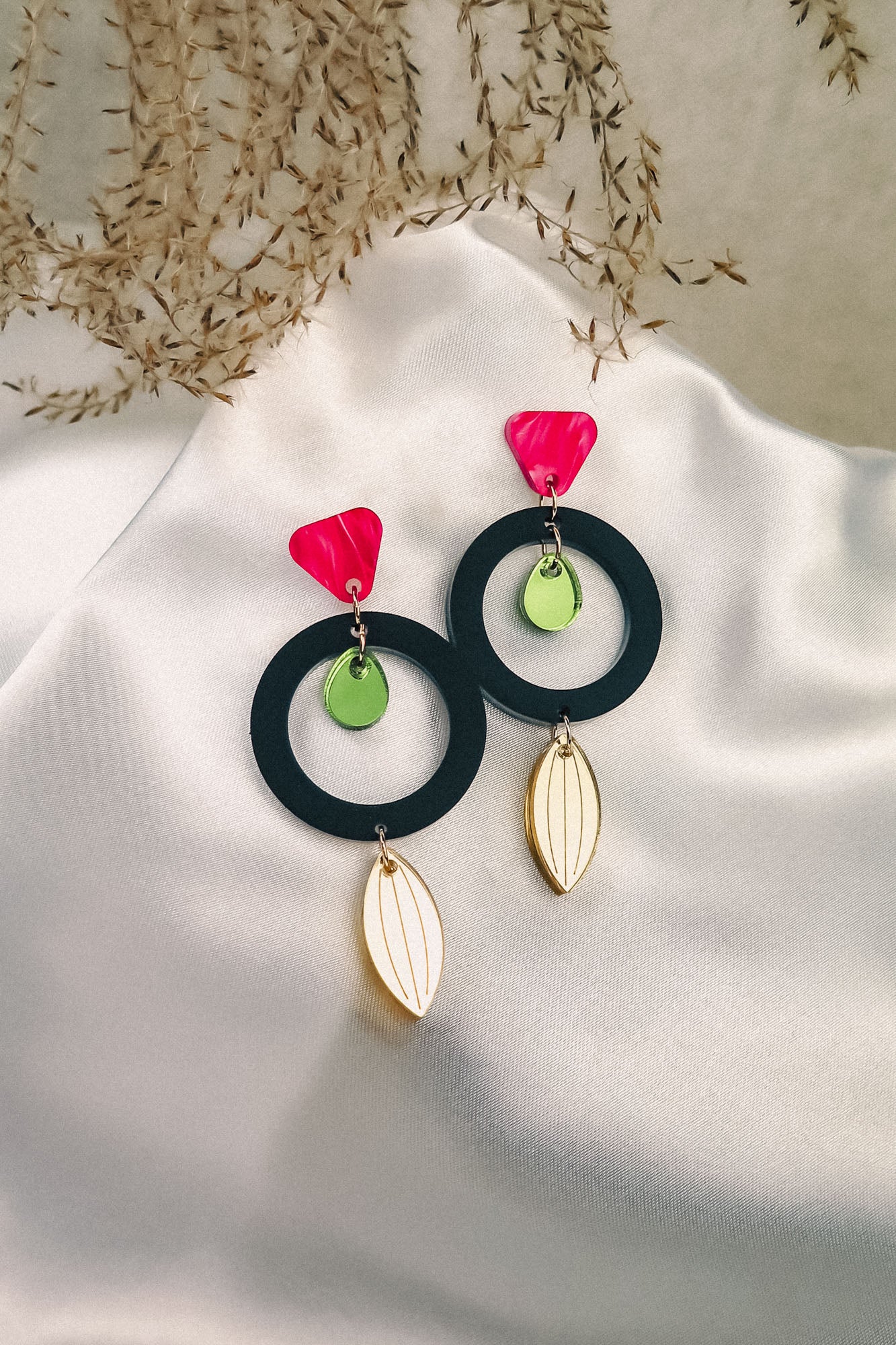 A pair of modern, colorful dangle earrings crafted from recycled acrylic, featuring pink triangle studs, green circular accents, black hoops, and gold leaf-shaped dangles displayed on white satin with dried foliage in the background.