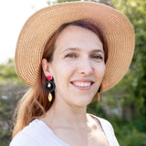 A woman with light brown hair wearing a wide-brimmed straw hat, artisan dangle earrings, and a white top smiles outdoors, surrounded by greenery and sunlight.