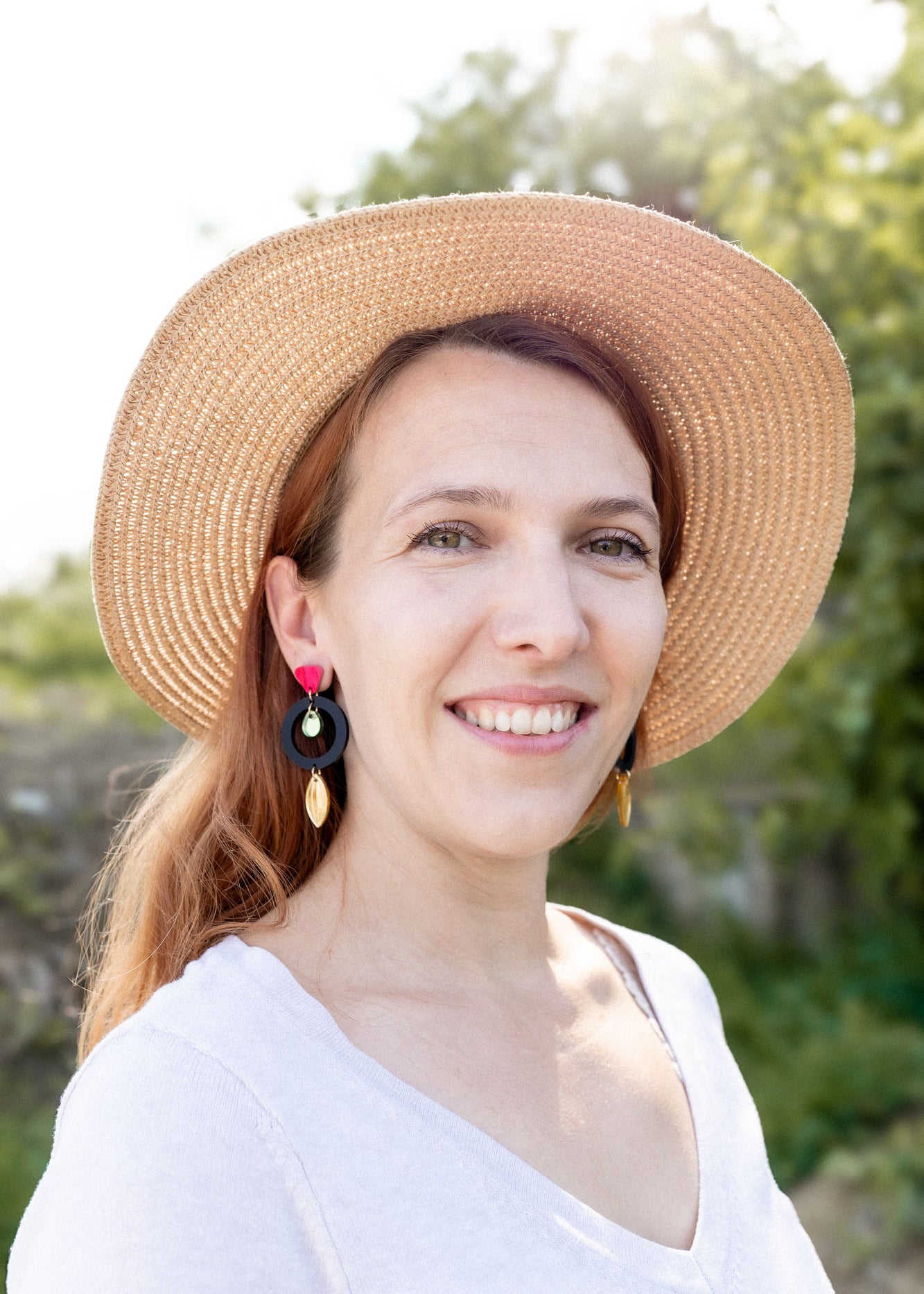 A woman with light brown hair wearing a wide-brimmed straw hat, artisan dangle earrings, and a white top smiles outdoors, surrounded by greenery and sunlight.