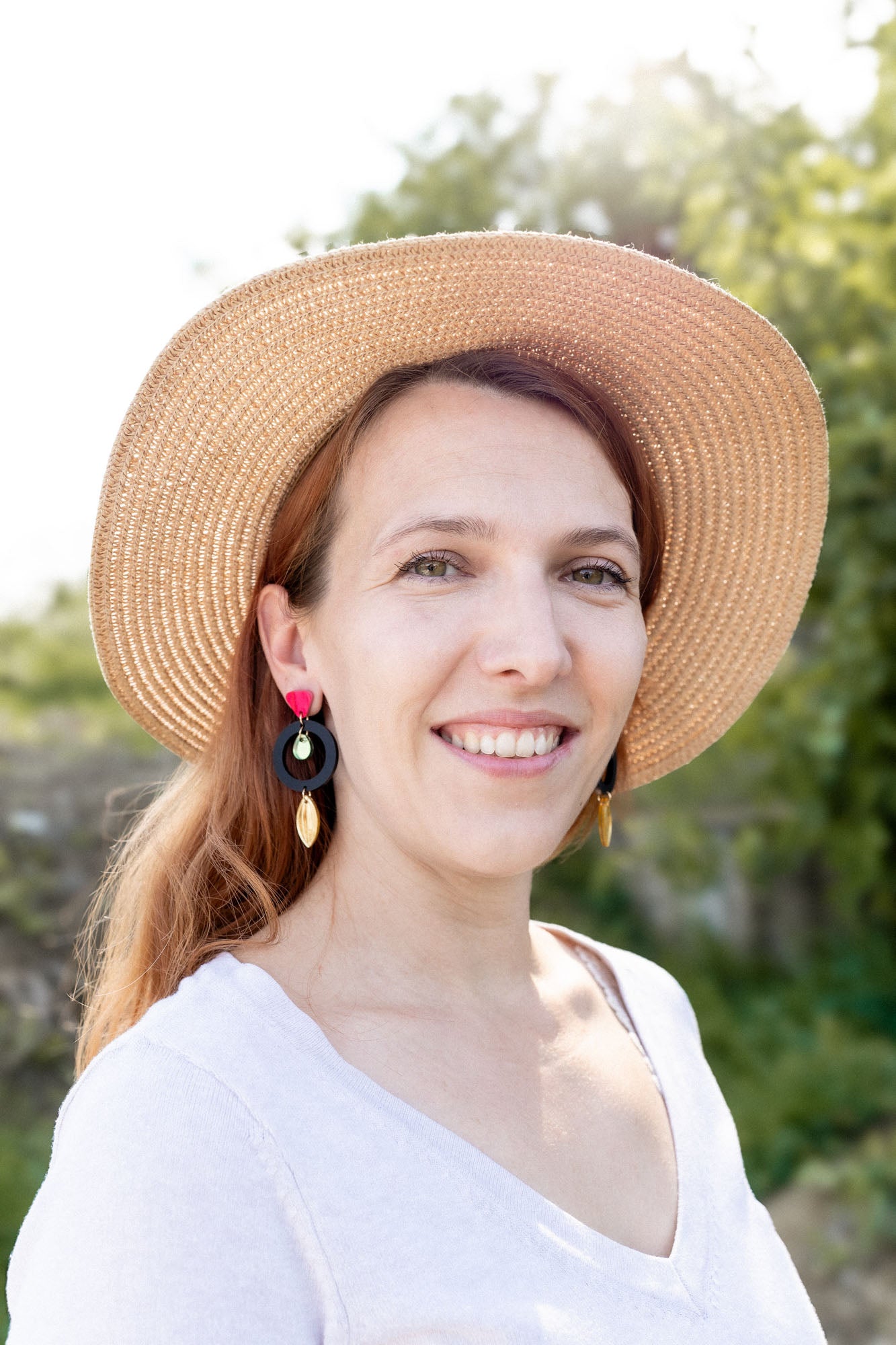A woman with light brown hair wearing a wide-brimmed straw hat, artisan dangle earrings, and a white top smiles outdoors, surrounded by greenery and sunlight.
