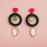 Pair of modern dangle earrings crafted from recycled acrylic, featuring black circular hoops, pink petal-shaped tops, small green discs, and gold leaf-shaped dangles, displayed on a soft pink background. Perfect artisan jewelry statement.