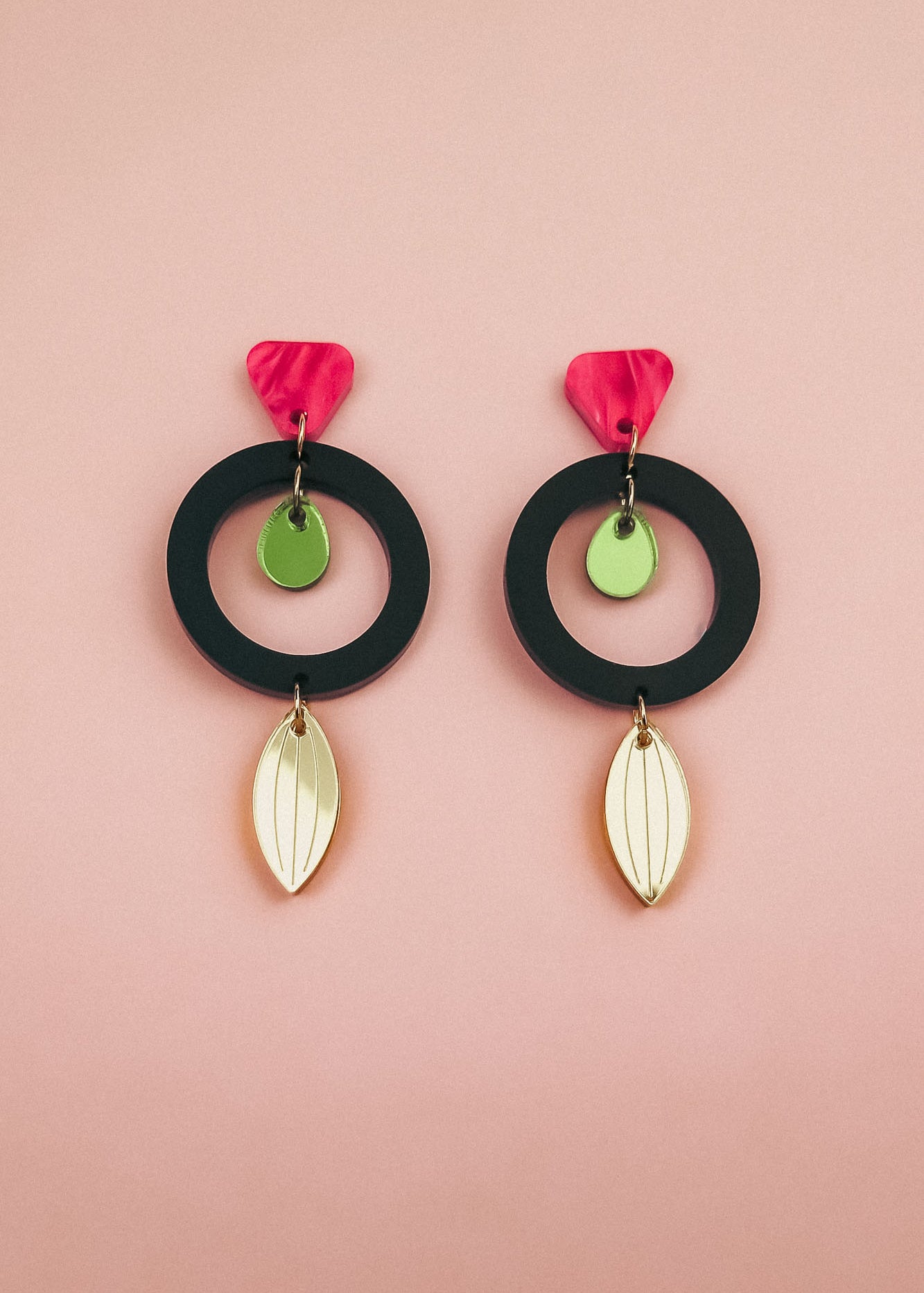 Pair of modern dangle earrings crafted from recycled acrylic, featuring black circular hoops, pink petal-shaped tops, small green discs, and gold leaf-shaped dangles, displayed on a soft pink background. Perfect artisan jewelry statement.