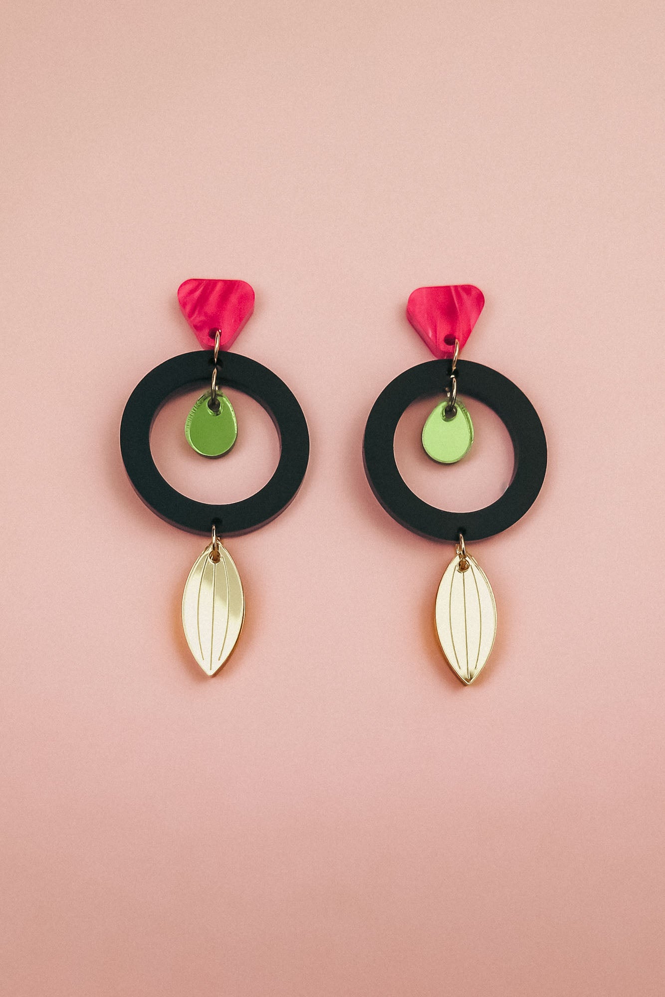 Pair of modern dangle earrings crafted from recycled acrylic, featuring black circular hoops, pink petal-shaped tops, small green discs, and gold leaf-shaped dangles, displayed on a soft pink background. Perfect artisan jewelry statement.