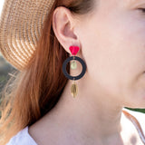 A woman in a straw hat and white shirt showcases artisan jewelry: dangle earrings crafted from recycled acrylic, featuring a black circle, gold shapes, and a red accent near the ear for a striking statement look.