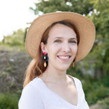 A woman wearing a wide-brimmed straw hat and artisan dangle earrings smiles outdoors. She has light brown hair and is dressed in a light-colored top, with greenery and trees in the blurred background.