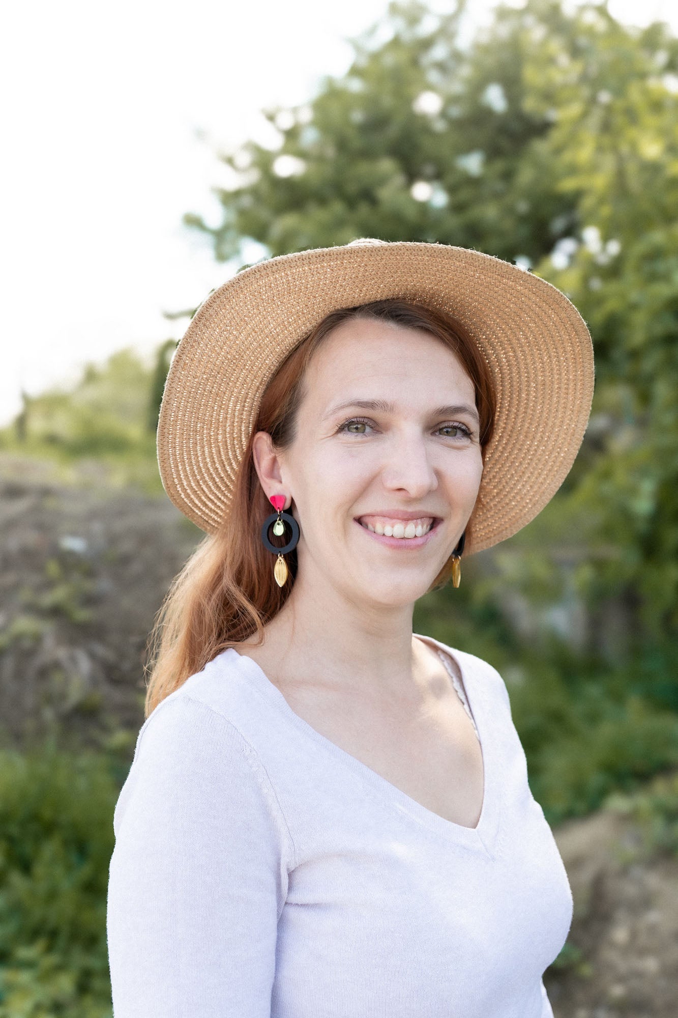 A woman wearing a wide-brimmed straw hat and artisan dangle earrings smiles outdoors. She has light brown hair and is dressed in a light-colored top, with greenery and trees in the blurred background.