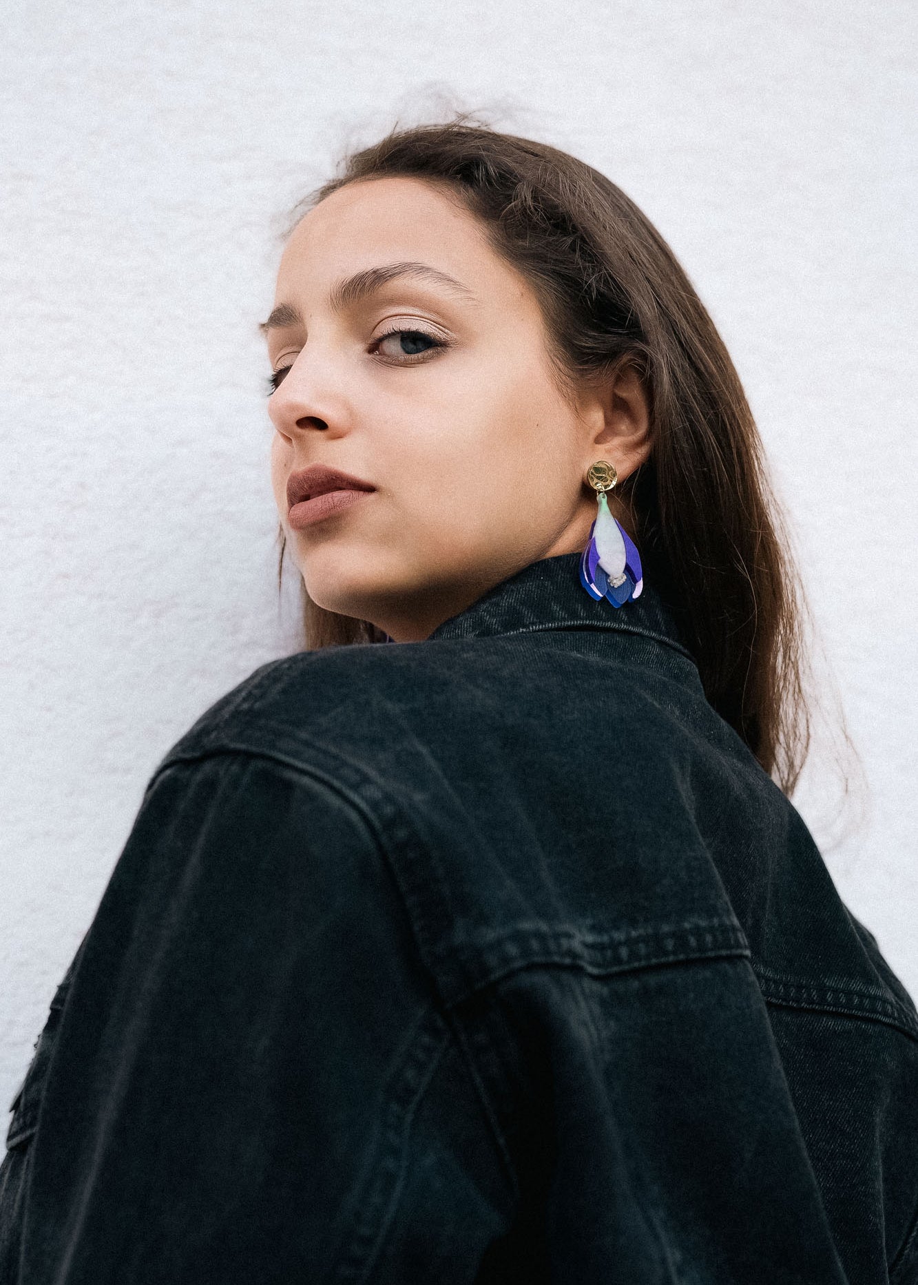 Woman with long brown hair wears a black denim jacket and handcrafted Crocus Flower Earrings, looking over her shoulder against a plain, light-colored background.