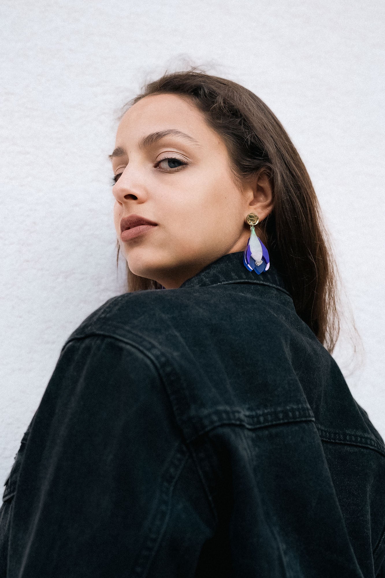 Woman with long brown hair wears a black denim jacket and handcrafted Crocus Flower Earrings, looking over her shoulder against a plain, light-colored background.
