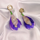 A pair of handcrafted Crocus Flower Earrings, resembling purple and pink blooms with gold circular studs, displayed on smooth white satin fabric—a stunning statement accessory.