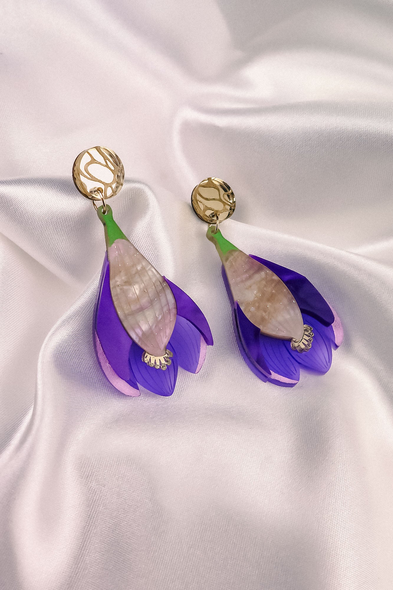 A pair of handcrafted Crocus Flower Earrings, resembling purple and pink blooms with gold circular studs, displayed on smooth white satin fabric—a stunning statement accessory.