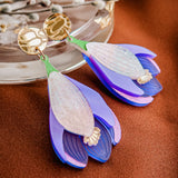 A pair of handcrafted Crocus Flower Earrings, shaped like purple and white blossoms with gold circular studs, rest on a brown surface near a glass dish and decorative twig—a stunning statement accessory.