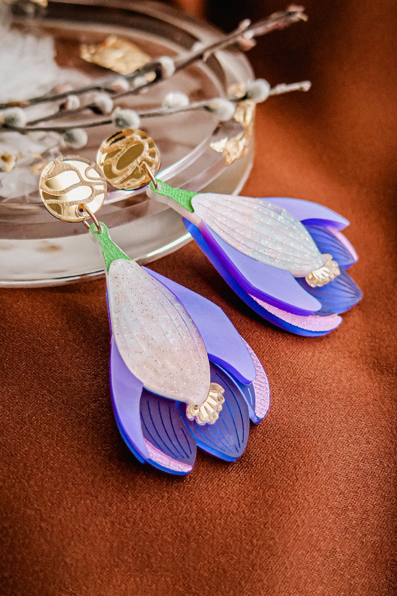 A pair of handcrafted Crocus Flower Earrings, shaped like purple and white blossoms with gold circular studs, rest on a brown surface near a glass dish and decorative twig—a stunning statement accessory.