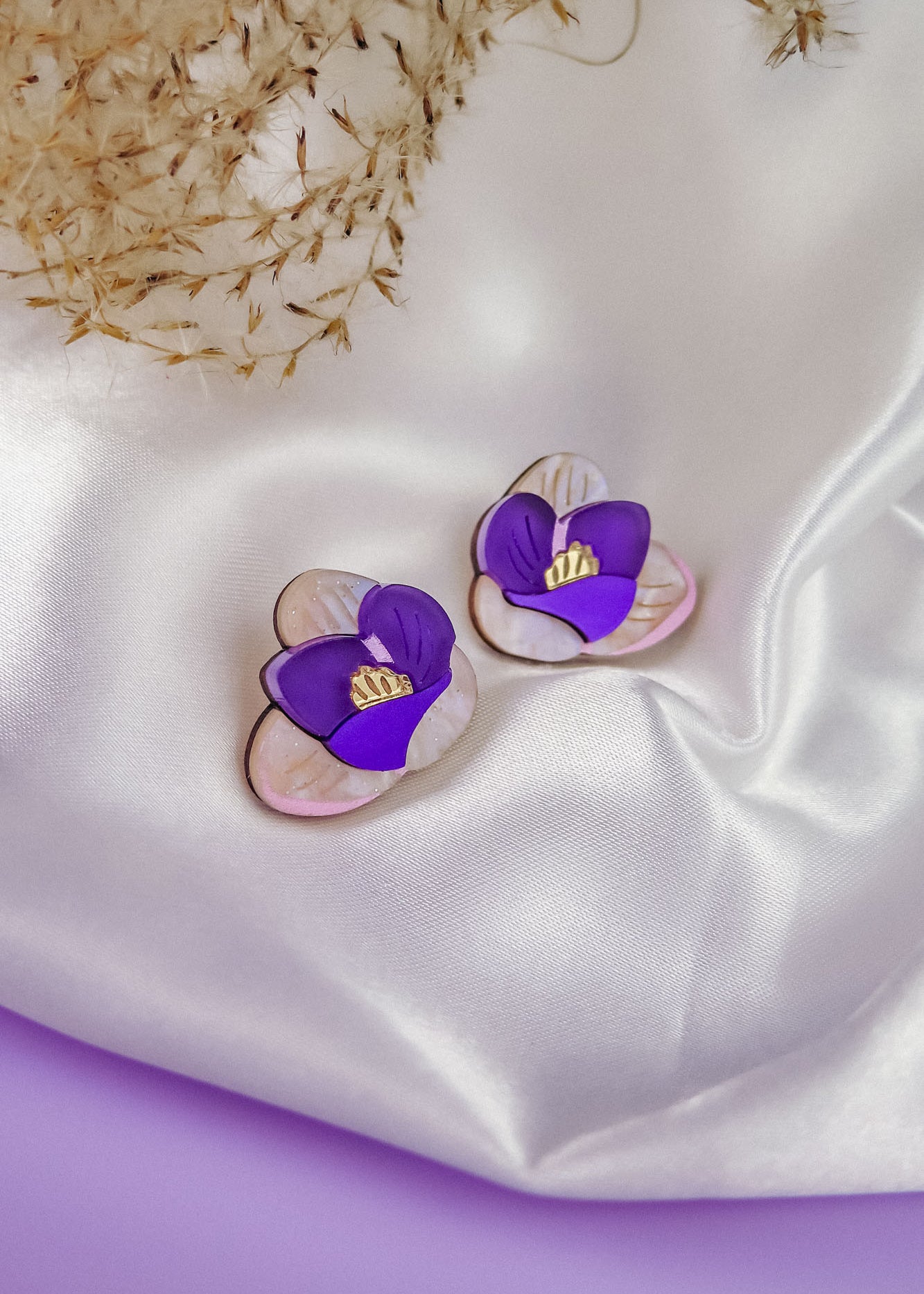 Crocus Stud Earrings shaped like hand-painted flowers with purple petals and yellow centers, displayed on white satin fabric with dried grasses in the background. These beautiful earrings are allergy-free.