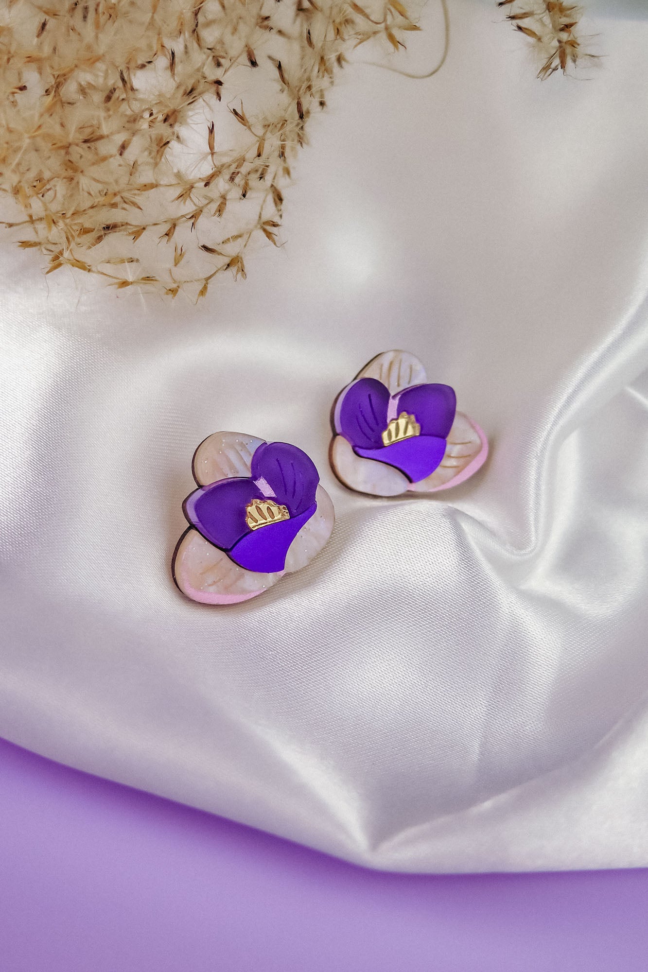 Crocus Stud Earrings shaped like hand-painted flowers with purple petals and yellow centers, displayed on white satin fabric with dried grasses in the background. These beautiful earrings are allergy-free.