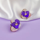 Two purple and white Crocus Stud Earrings, featuring hand-painted flowers, are displayed on a smooth lavender surface, with a white satin cloth and dried flowers in the background.