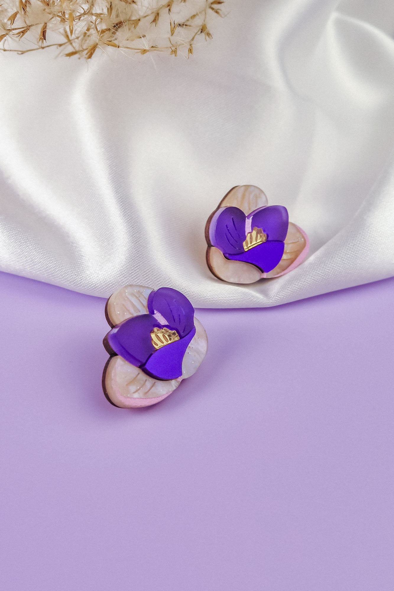 Two purple and white Crocus Stud Earrings, featuring hand-painted flowers, are displayed on a smooth lavender surface, with a white satin cloth and dried flowers in the background.