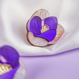 A decorative brooch shaped like a purple and cream hand-painted flower with gold details rests on silky white fabric. Another similar brooch is partially visible in the foreground.