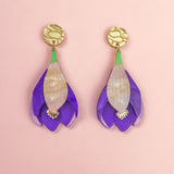 A pair of handcrafted Crocus Flower Earrings featuring purple petals with cream-colored centers and gold circular studs, displayed on a soft pink background—a stunning statement accessory.