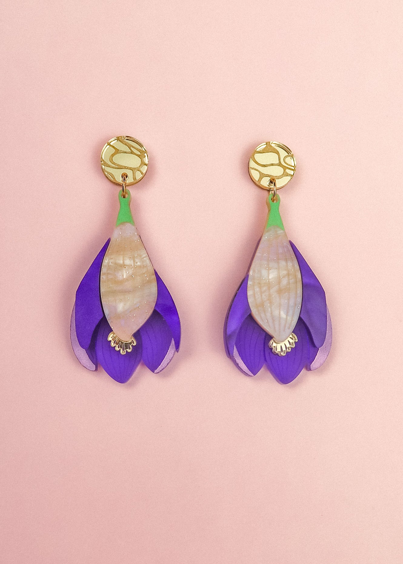 A pair of handcrafted Crocus Flower Earrings featuring purple petals with cream-colored centers and gold circular studs, displayed on a soft pink background—a stunning statement accessory.