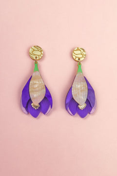 A pair of handcrafted Crocus Flower Earrings featuring purple petals with cream-colored centers and gold circular studs, displayed on a soft pink background—a stunning statement accessory.