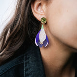 Close-up of a person wearing Crocus Flower Earrings—a handcrafted, statement accessory shaped like a flower petal in purple, white, and gold. The person has long brown hair and is dressed in a dark-colored top.