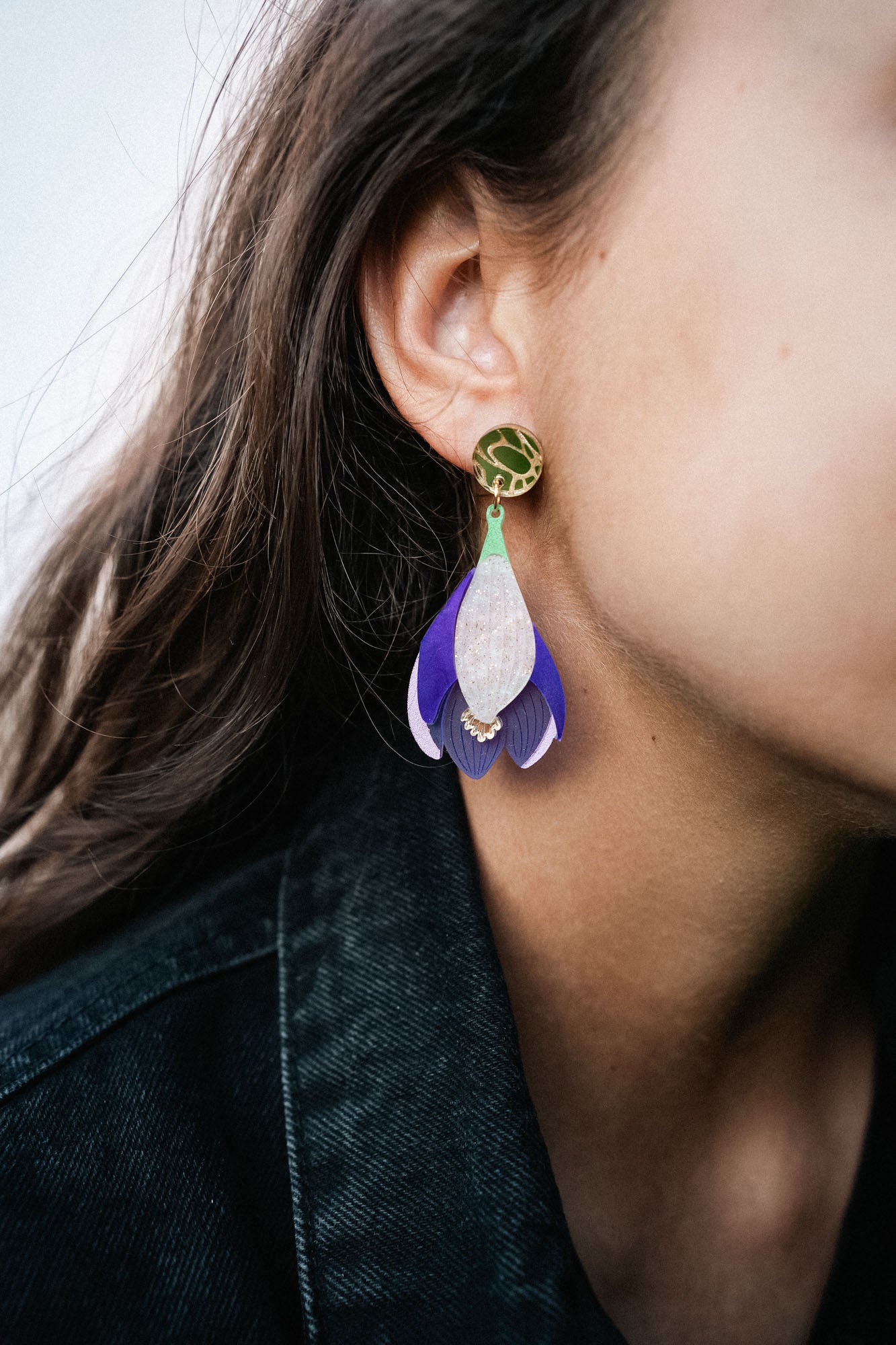 Close-up of a person wearing Crocus Flower Earrings—a handcrafted, statement accessory shaped like a flower petal in purple, white, and gold. The person has long brown hair and is dressed in a dark-colored top.