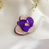 A purple and cream flower-shaped brooch, reminiscent of a Purple Crocus Flower Lapel Pin with gold detailing, rests on smooth white satin fabric, surrounded by delicate dried grass—perfect for nature lovers.