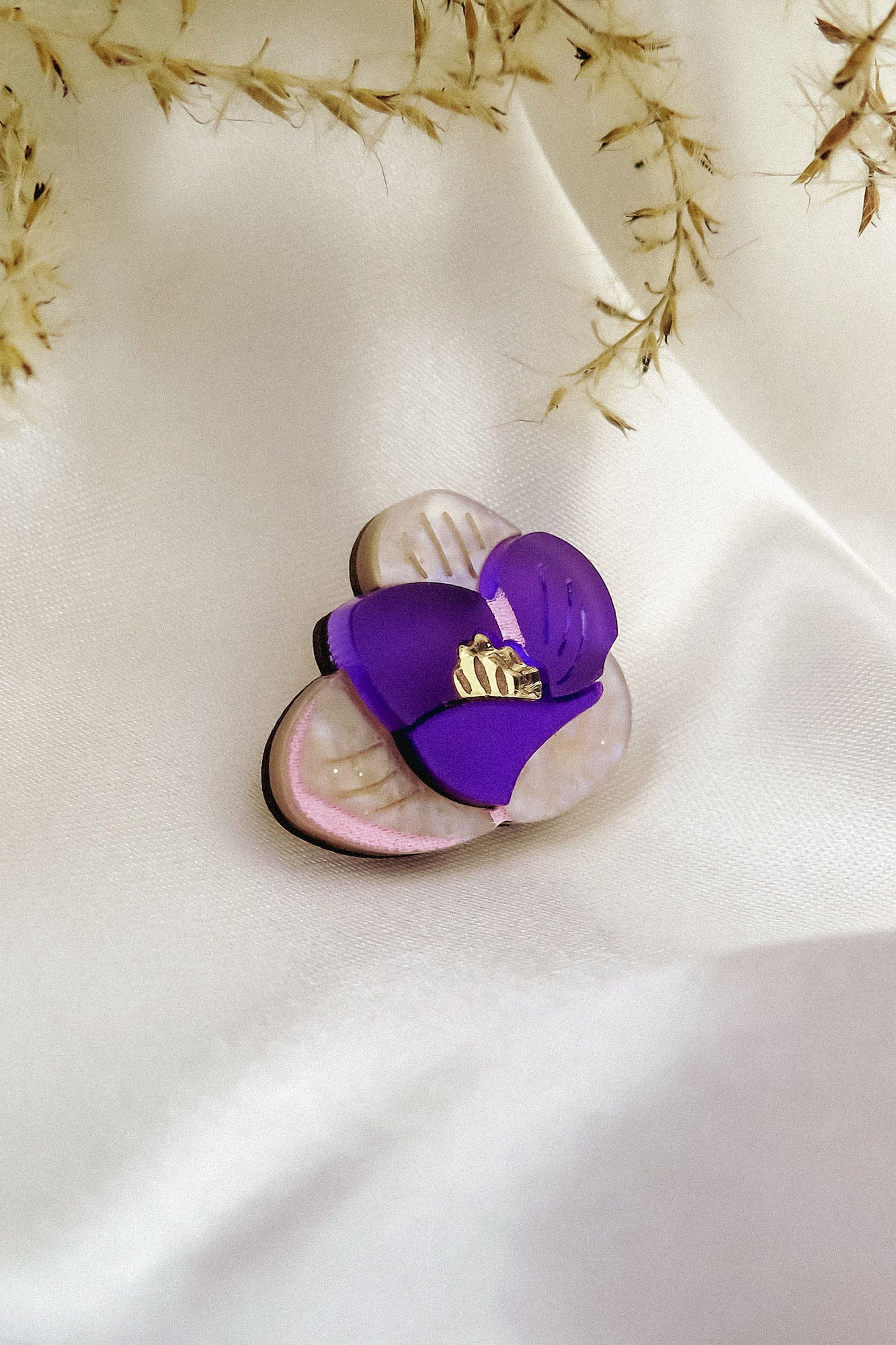 A purple and cream flower-shaped brooch, reminiscent of a Purple Crocus Flower Lapel Pin with gold detailing, rests on smooth white satin fabric, surrounded by delicate dried grass—perfect for nature lovers.