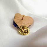A handcrafted gold lapel pin with an embossed hand design rests on a wooden heart-shaped base, placed on a smooth white fabric background—an ideal gift for nature lovers.