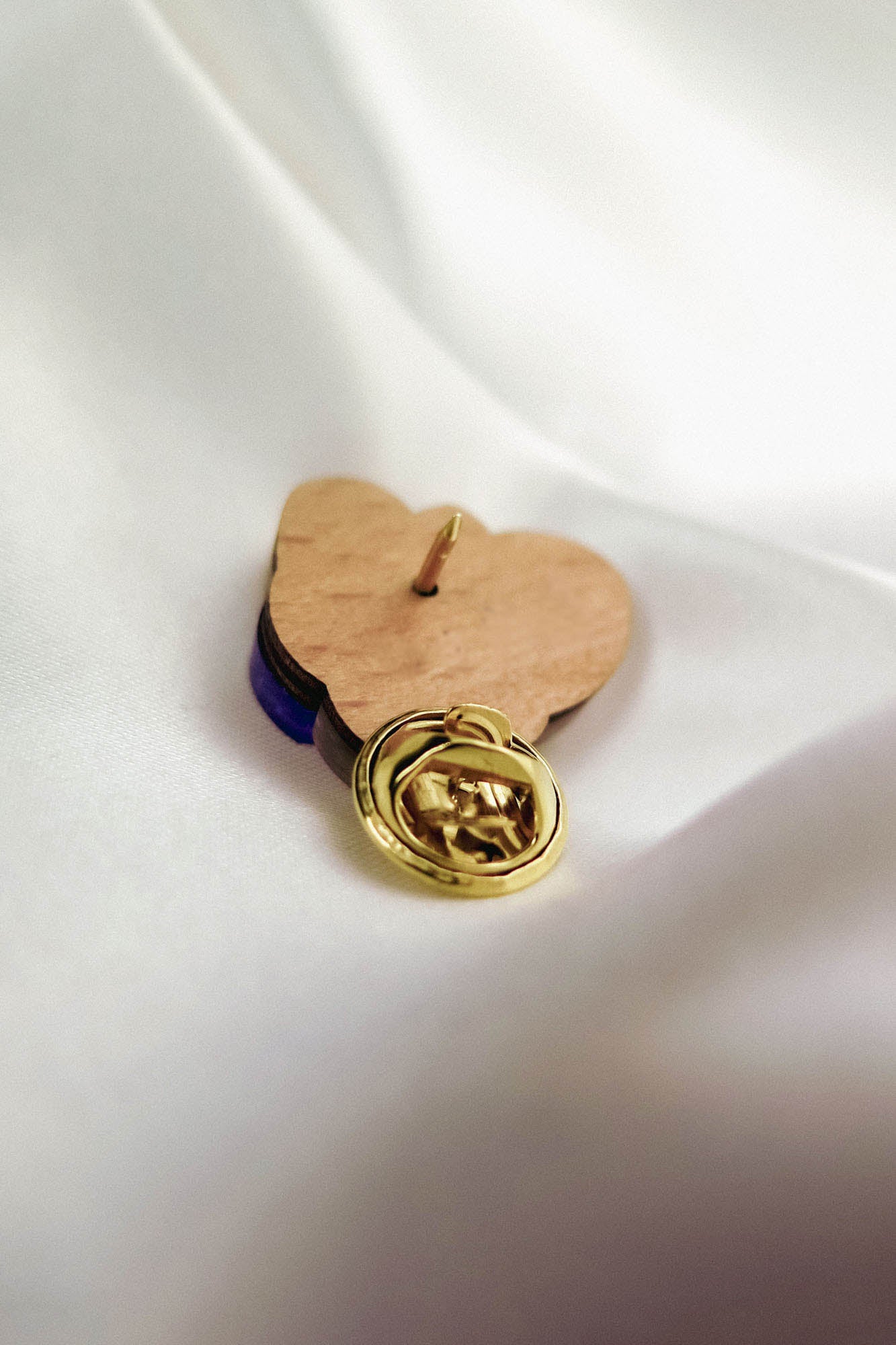A handcrafted gold lapel pin with an embossed hand design rests on a wooden heart-shaped base, placed on a smooth white fabric background—an ideal gift for nature lovers.