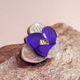 A close-up of a handcrafted Purple Crocus Flower Lapel Pin with purple and white petals and a gold center, resting on a textured, metallic-looking surface—perfect for nature lovers.