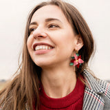 A woman with long brown hair smiles while wearing a red sweater, a checkered coat, and Poinsettia Statement Hoops, adding a touch of festive jewelry. The background is outdoors with a cloudy sky.