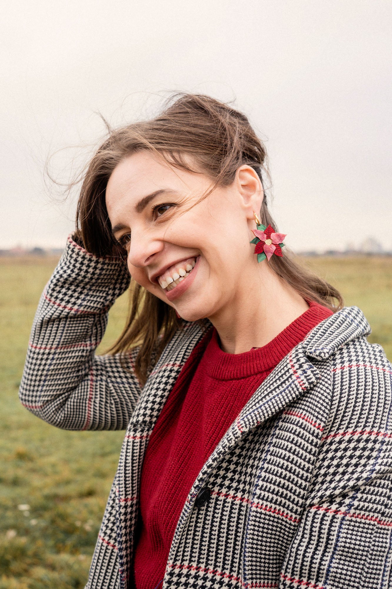 A smiling woman with brown hair stands outdoors in a grassy field, wearing a red sweater, a plaid coat, and festive Poinsettia Statement Hoops. She holds her hair back with one hand on a cloudy day, adding a touch of holiday earrings to her look.