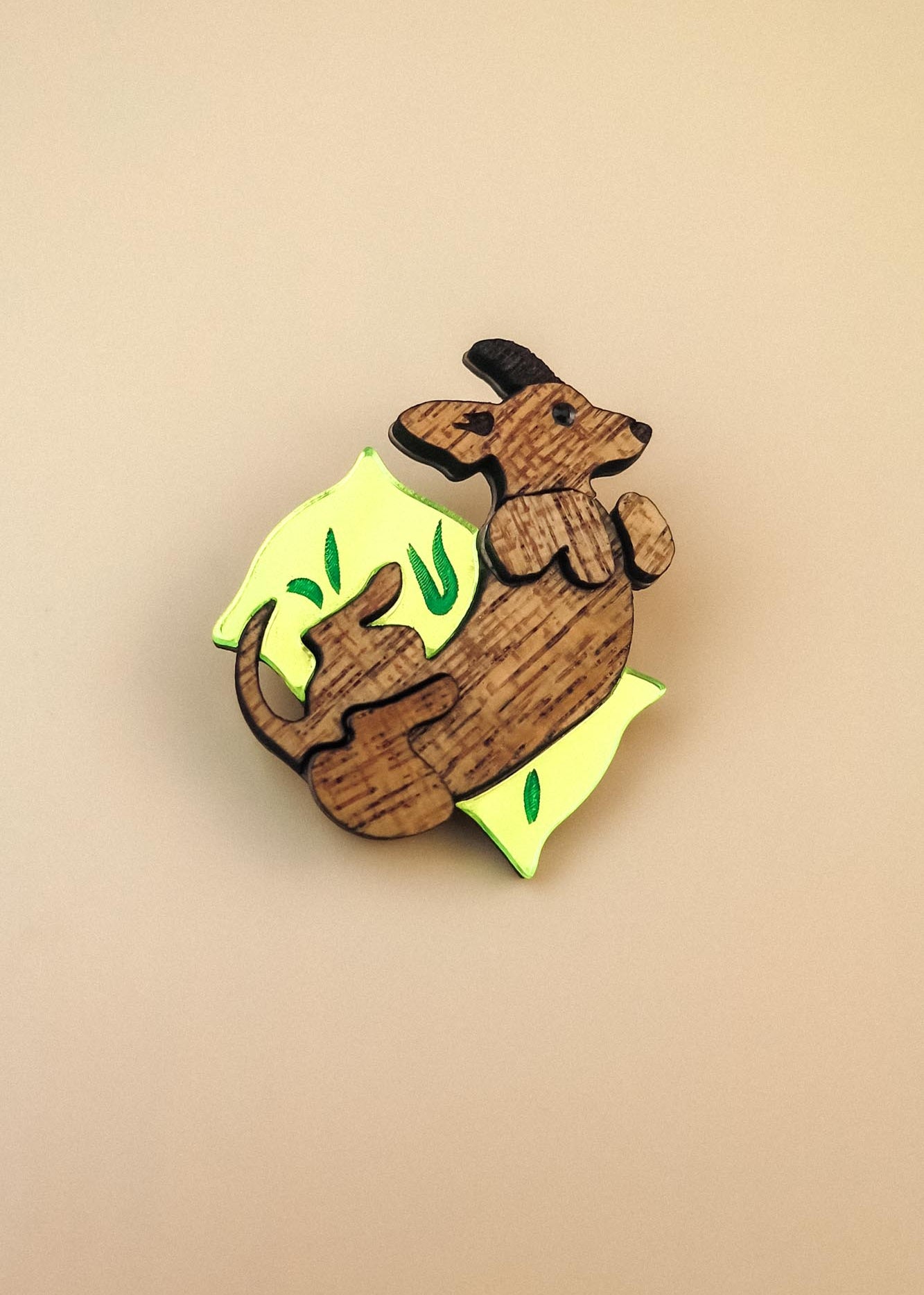A playful brooch shaped like a dachshund lies on a plain beige background. With engraved details and bright yellow and green leaf-like accents, this wooden piece adds whimsy to any dog accessory collection.